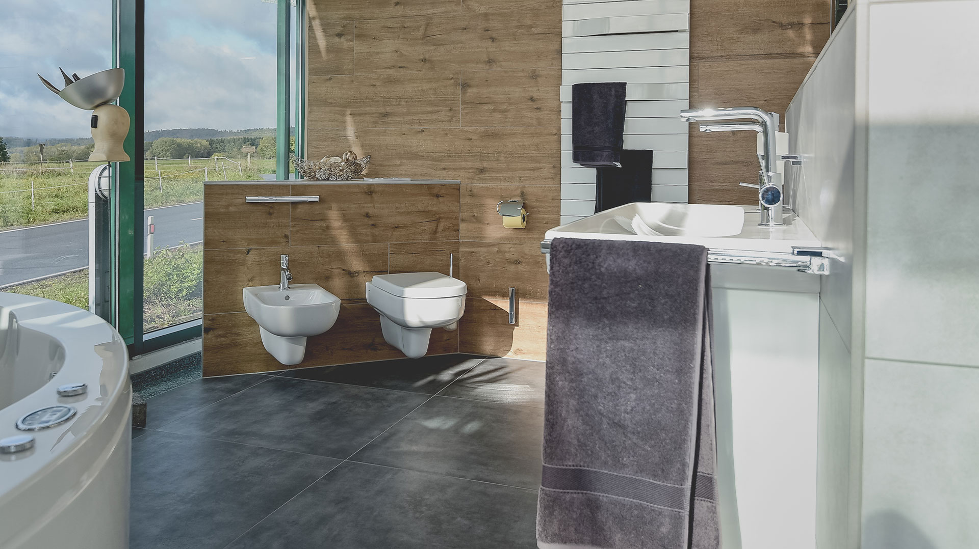 Sonnenlicht fällt in ein modernes Badezimmer mit großem Fenster, Holzverkleidung, wandhängendem WC und Bidet sowie weißem Waschtisch mit Chromarmatur.